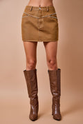 A model's lower body wearing a brown denim mini skirt with silver rhinestone embellishments on the front waistband, paired with brown knee-high textured boots, against a solid beige studio background.