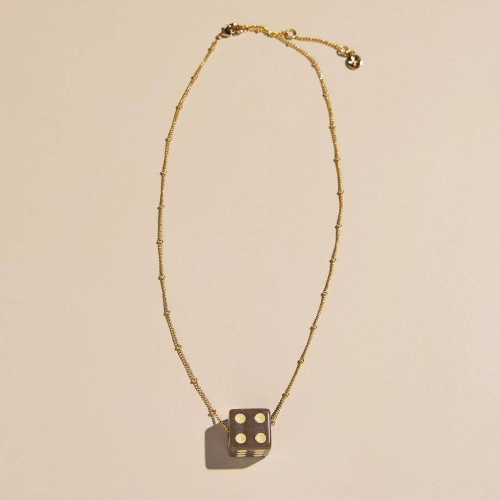A gold beaded chain necklace with a dark metallic dice pendant showing four white dots, displayed on a warm beige background.