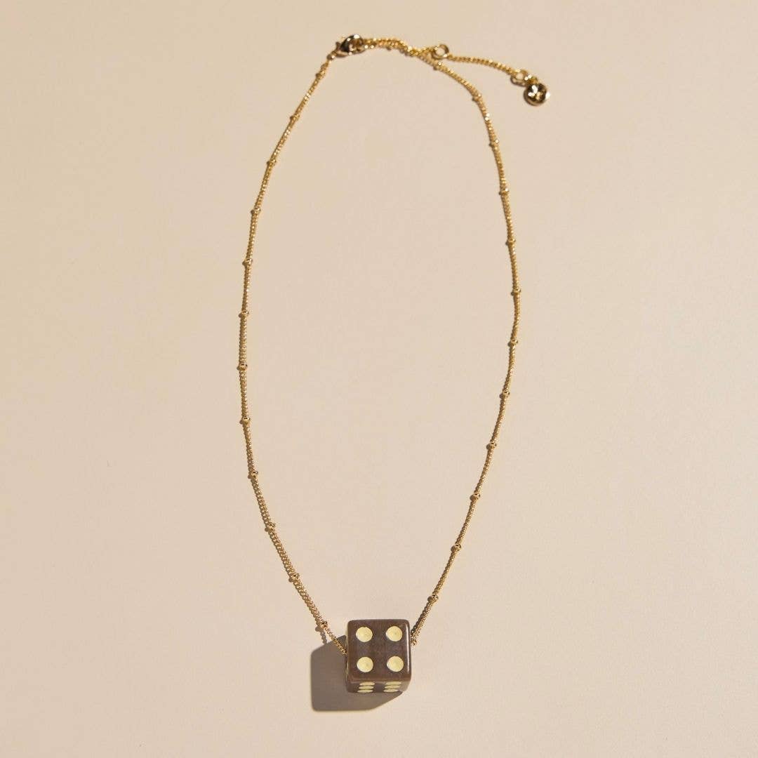 A gold beaded chain necklace with a dark metallic dice pendant showing four white dots, displayed on a warm beige background.
