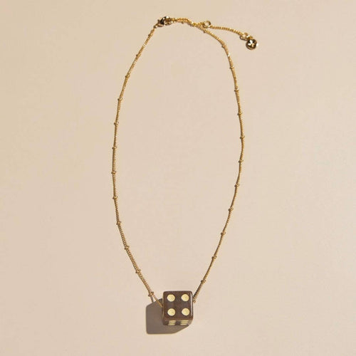 A gold beaded chain necklace with a dark metallic dice pendant showing four white dots, displayed on a warm beige background.