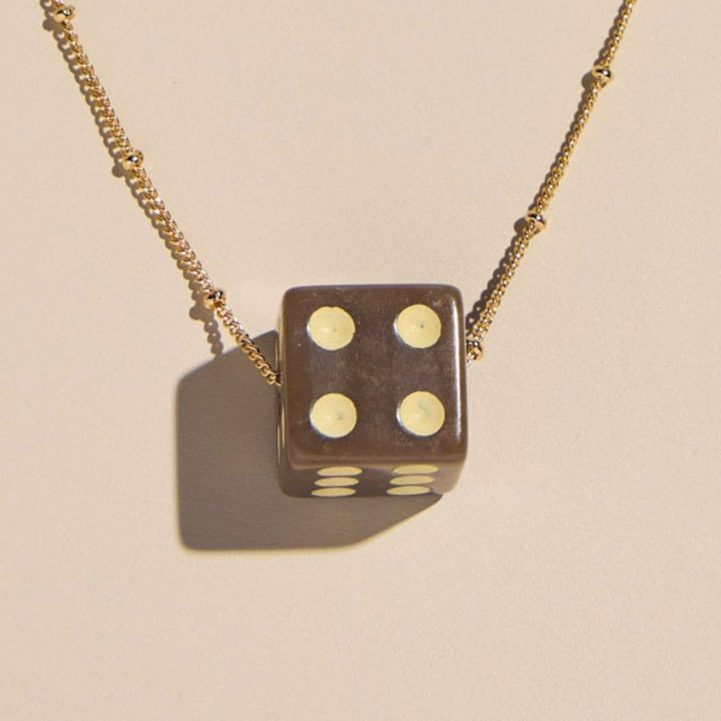 A gold chain necklace featuring a brown die-shaped pendant with cream-colored pips, shown against a warm beige background with a soft shadow.