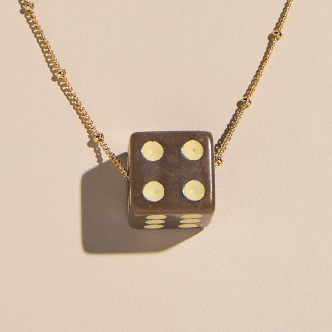 A gold chain necklace featuring a brown die-shaped pendant with cream-colored pips, shown against a warm beige background with a soft shadow.