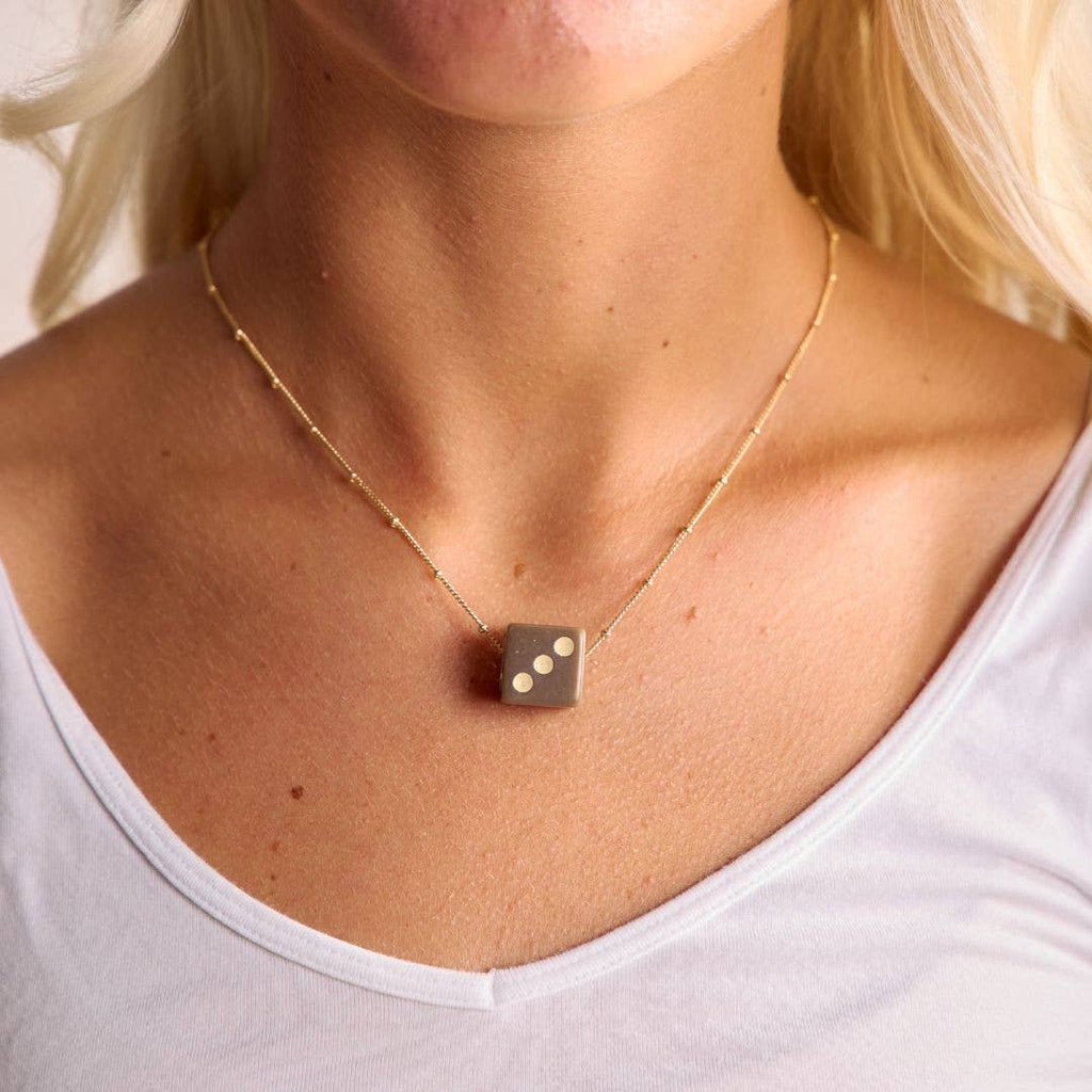 A close-up of a woman wearing a delicate gold chain necklace with a square dark gray pendant featuring three white dots, styled with a white tank top.