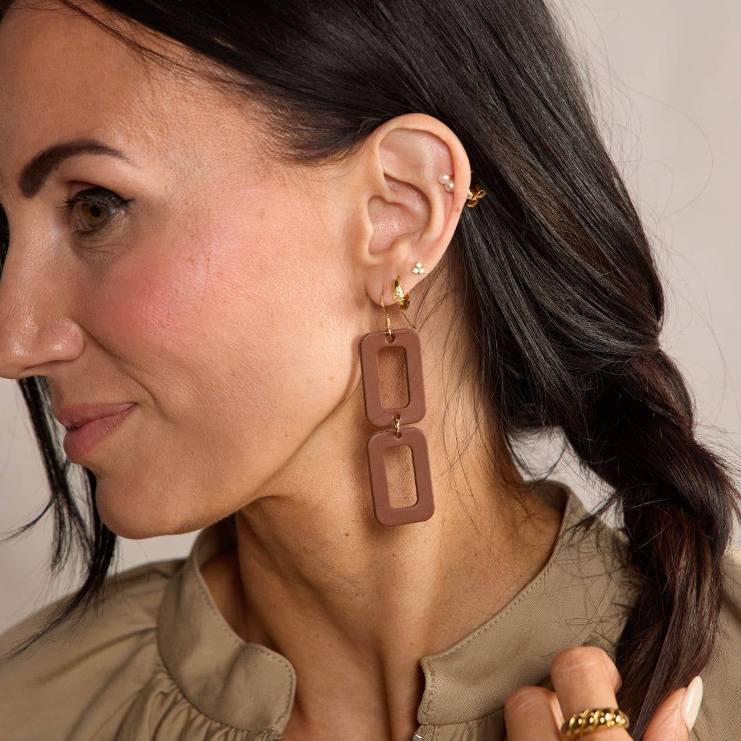 A close-up of a woman wearing a pair of large, double-link geometric drop earrings in a matte dusty rose-brown color. The earrings feature a small gold hoop at the top and are shown against her ear and neck.