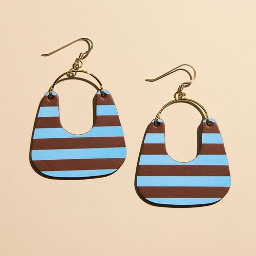 A pair of drop earrings with gold-tone fishhook wires and circular hoops, each holding a teardrop-shaped charm with alternating brown and light blue horizontal stripes, shown on a warm beige background.