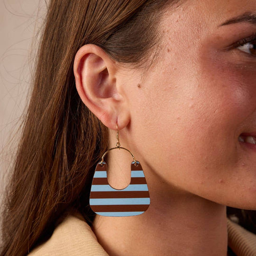 A close-up of a woman wearing a large teardrop-shaped drop earring with horizontal brown and light blue stripes and a gold-tone hoop.