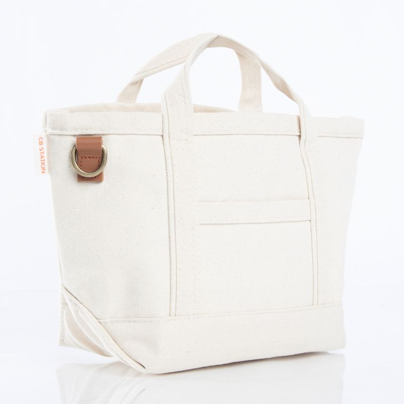 An off-white canvas tote bag with dual top handles, a front pocket, and a brown leather strap detail with a brass ring on the side, set against a plain white background.