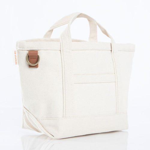 An off-white canvas tote bag with dual top handles, a front pocket, and a brown leather strap detail with a brass ring on the side, set against a plain white background.
