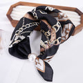 A black silk scarf with a brown and beige chain-link pattern and a white pearl accent, draped over a wooden jewelry display box on a white surface.