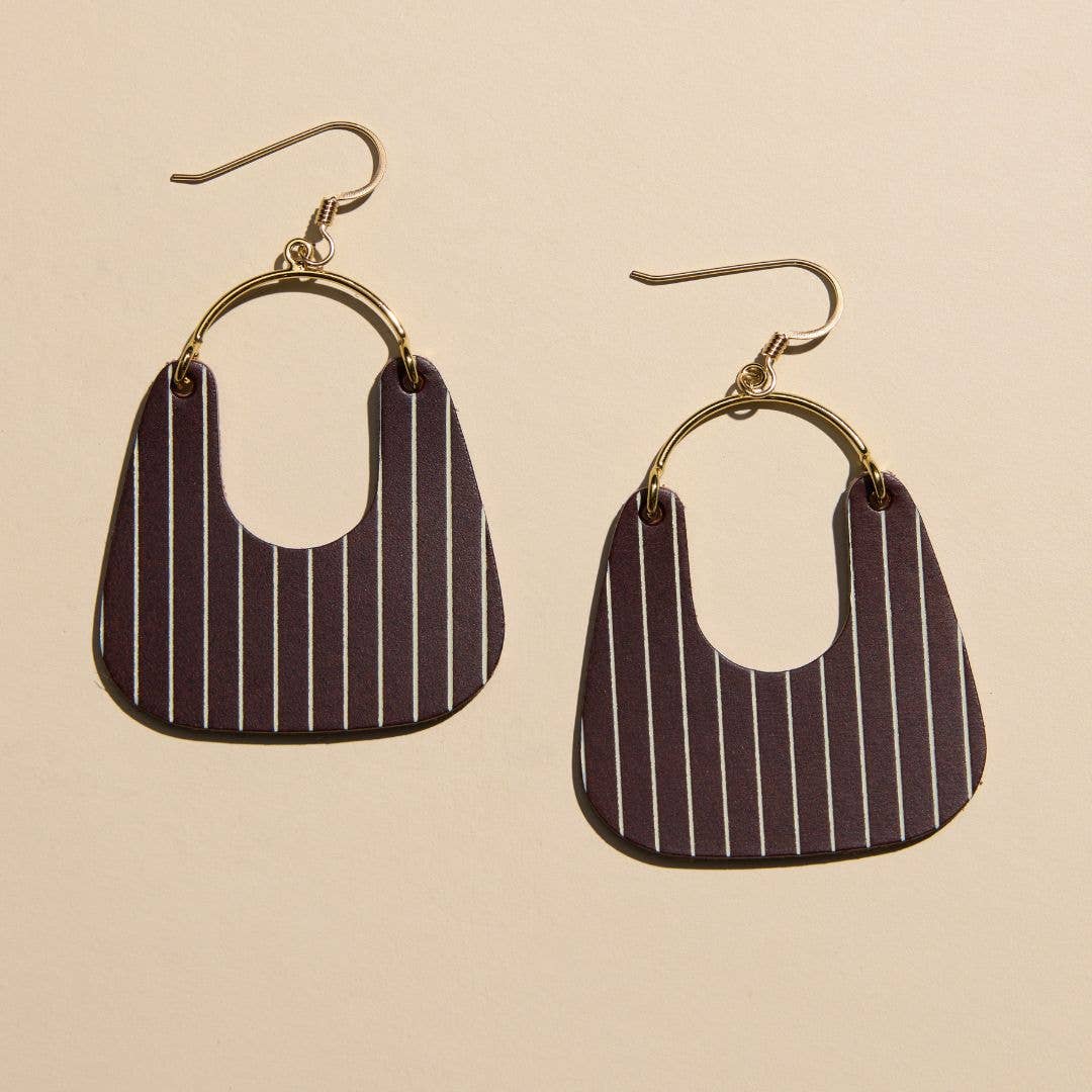 A pair of drop earrings with gold-tone fishhook wires and circular hoops connected to shield-shaped pinstriped charms in dark purple and white, shown on a light beige background.