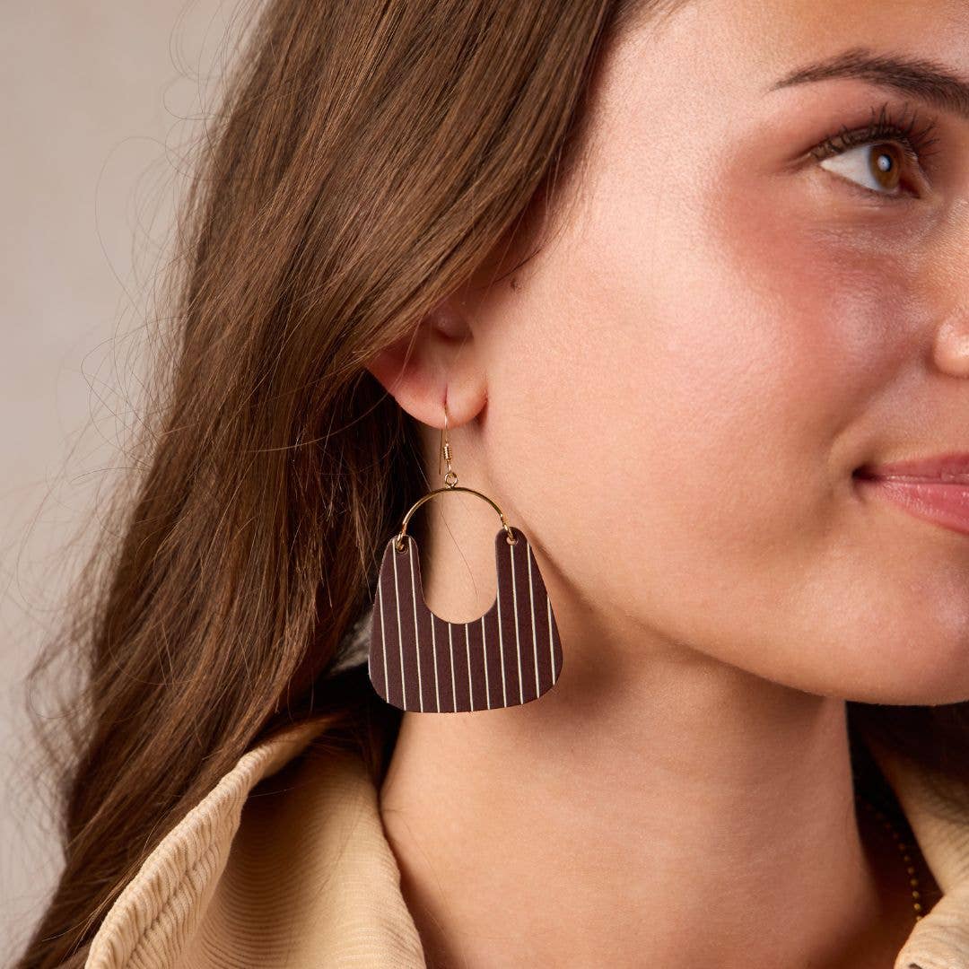 A close-up of a woman wearing a large, geometric brown dangle earring with thin white vertical stripes and a gold-tone hoop.