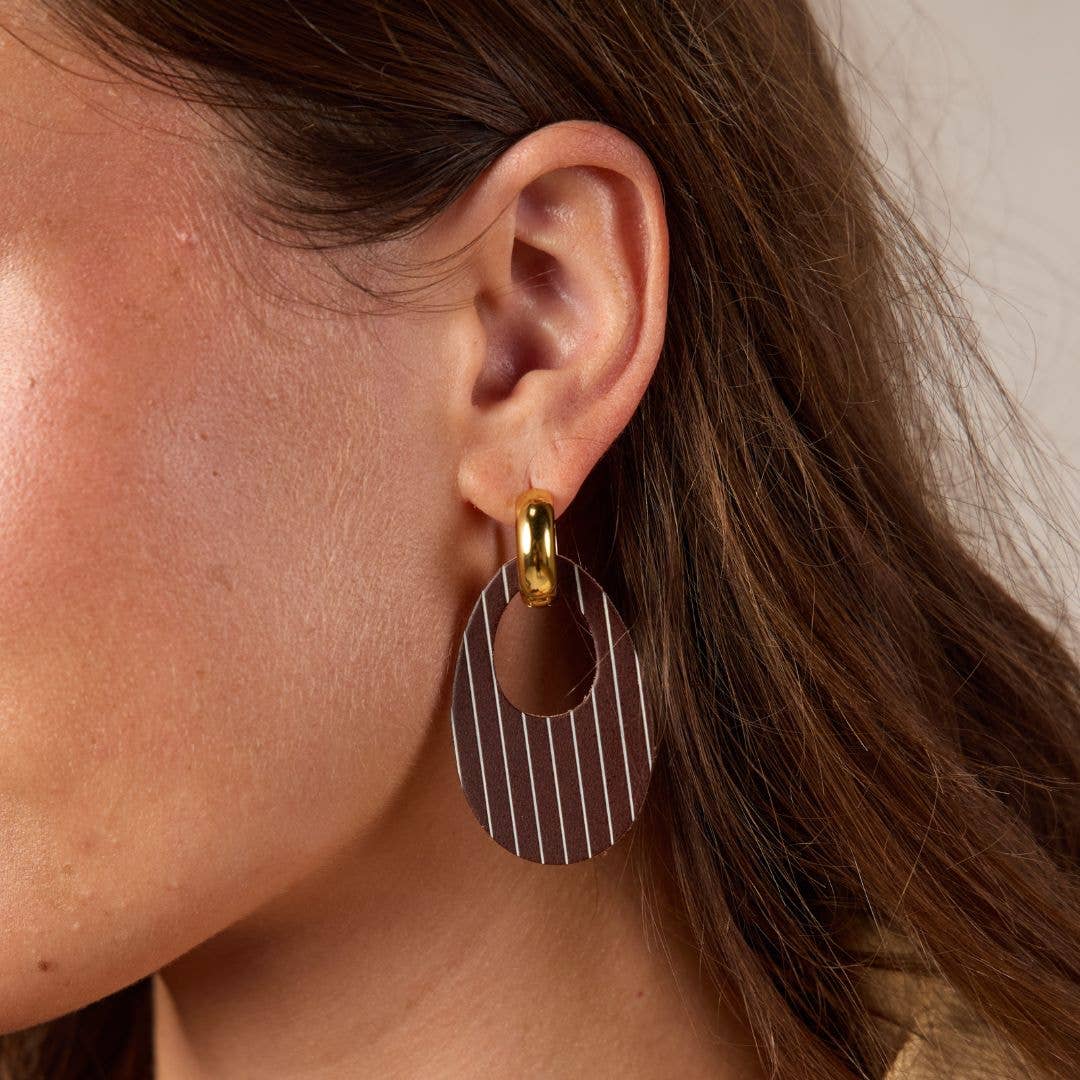 A close-up of a model's ear wearing a modern dangle earring. The earring features a gold-tone hoop hook attached to a large, flat circular drop with thin vertical white stripes on a dark brown background.