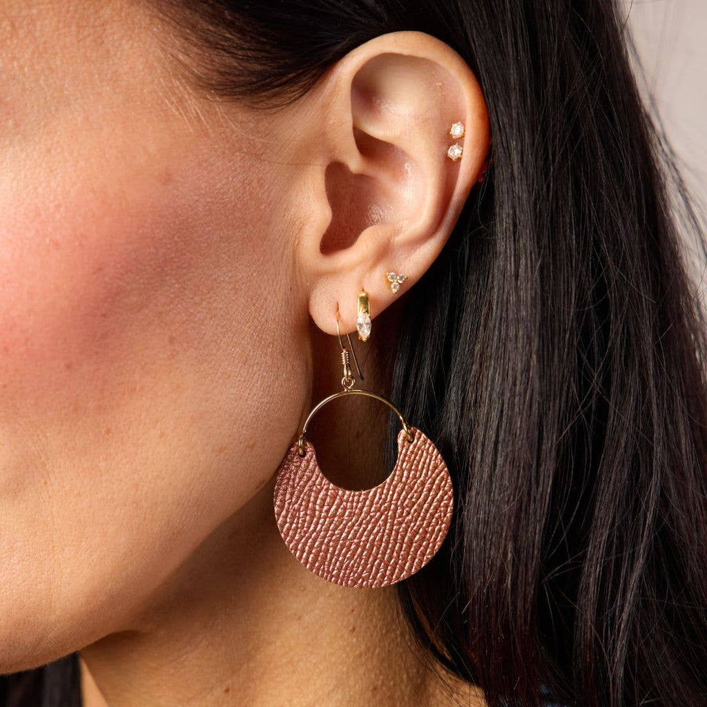 A close-up of a model's ear wearing a large textured rose gold crescent hoop earring with a gold hook, alongside smaller gold ear piercings.