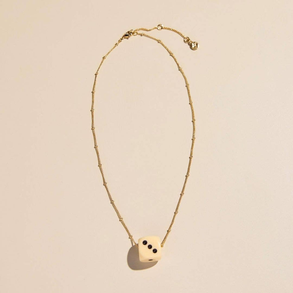 A delicate gold chain necklace featuring a cream-colored dice pendant with black pips, displayed on a warm beige background.