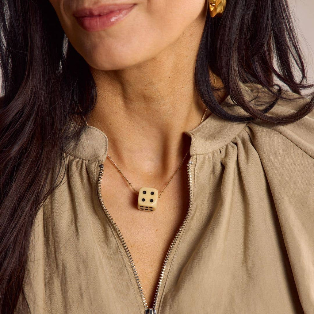 A close-up of a woman wearing a gold-tone necklace with a small dice pendant, paired with a beige zip-up top.