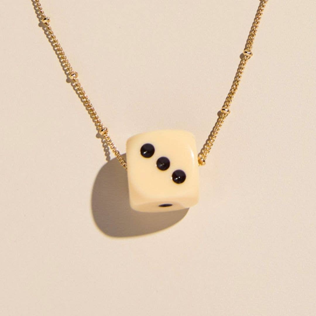 A gold chain necklace featuring a cream-colored die-shaped pendant with three black pips, shown against a warm beige background with a soft shadow.
