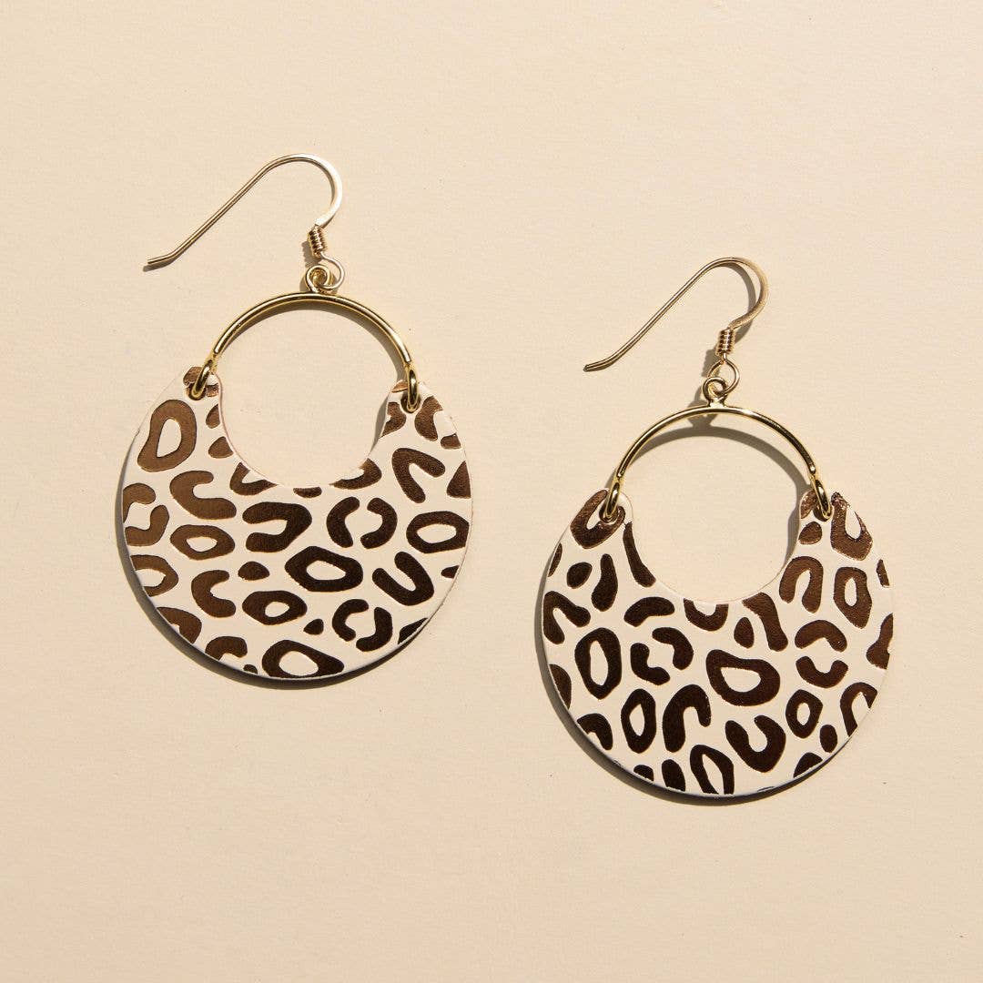 A pair of gold-tone hoop earrings with crescent-shaped dangles featuring a brown and beige leopard print pattern, shown on a warm beige background.