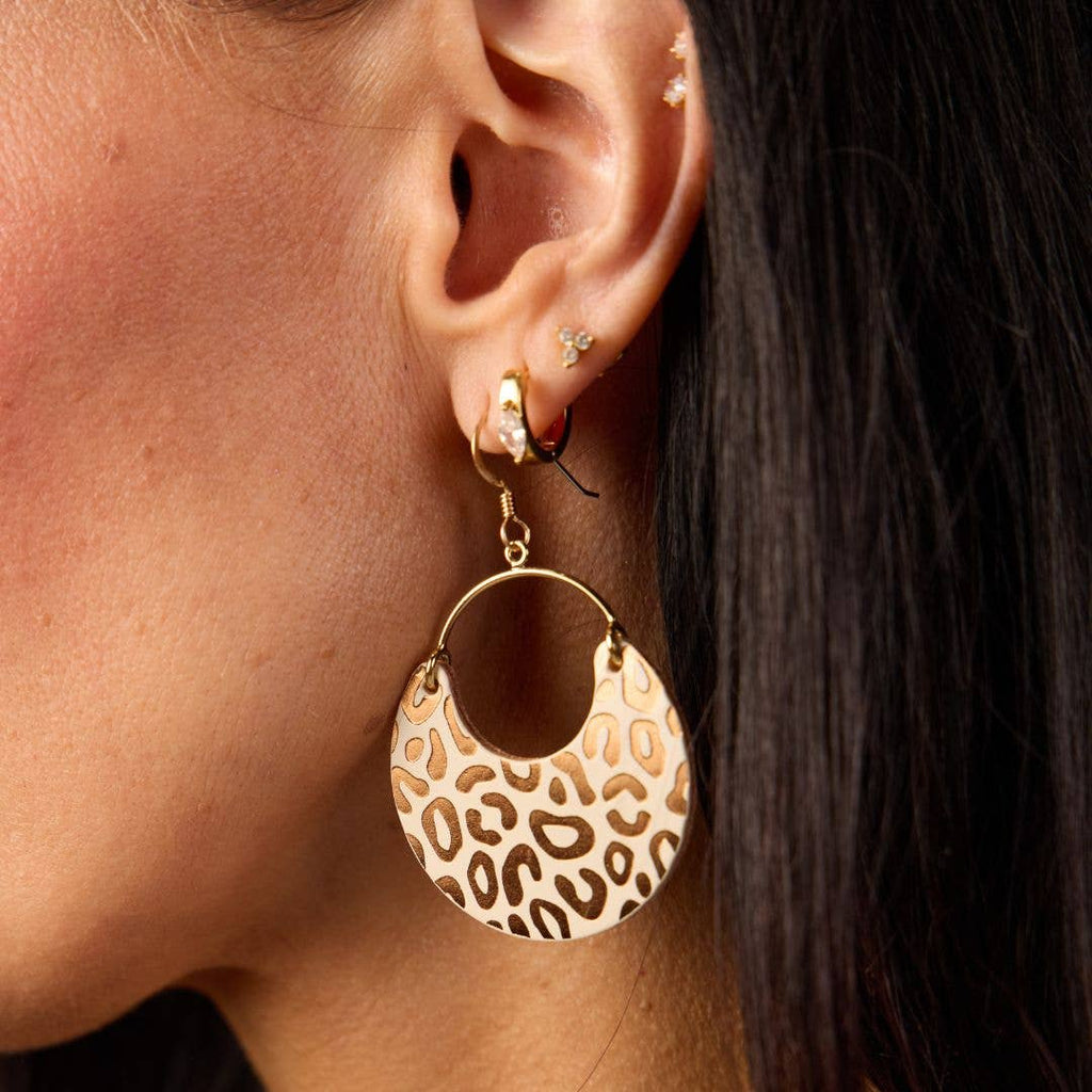 A close-up of a model's ear wearing a large gold hoop earring with a crescent-shaped dangle featuring a brown and beige leopard print pattern.