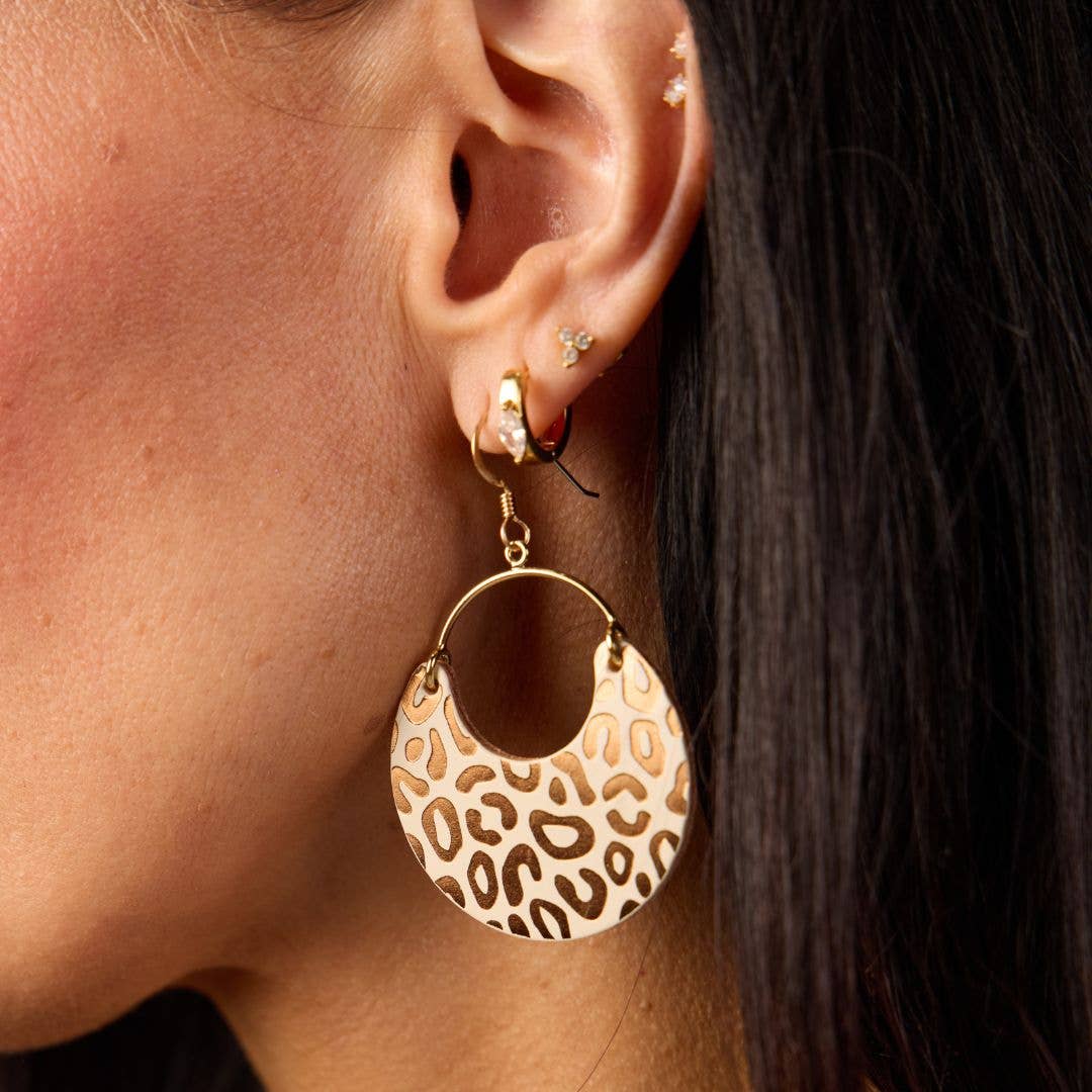 A close-up of a model's ear wearing a large gold hoop earring with a crescent-shaped dangle featuring a brown and beige leopard print pattern.