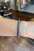 A close-up of a person's wrist wearing a beaded bracelet with bright turquoise-blue round beads separated by thin red spacer strands, photographed in a boutique setting with a blurred wooden floor and clothing rack in the background.