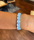 A close-up of a person's wrist wearing a beaded stretch bracelet made of light blue marbled beads, with a blurred boutique interior in the background.