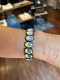 A close-up of a person's wrist wearing a dark brown elastic bracelet with seven oval iridescent cabochon gemstones that shimmer with rainbow colors, set against a blurred indoor background.