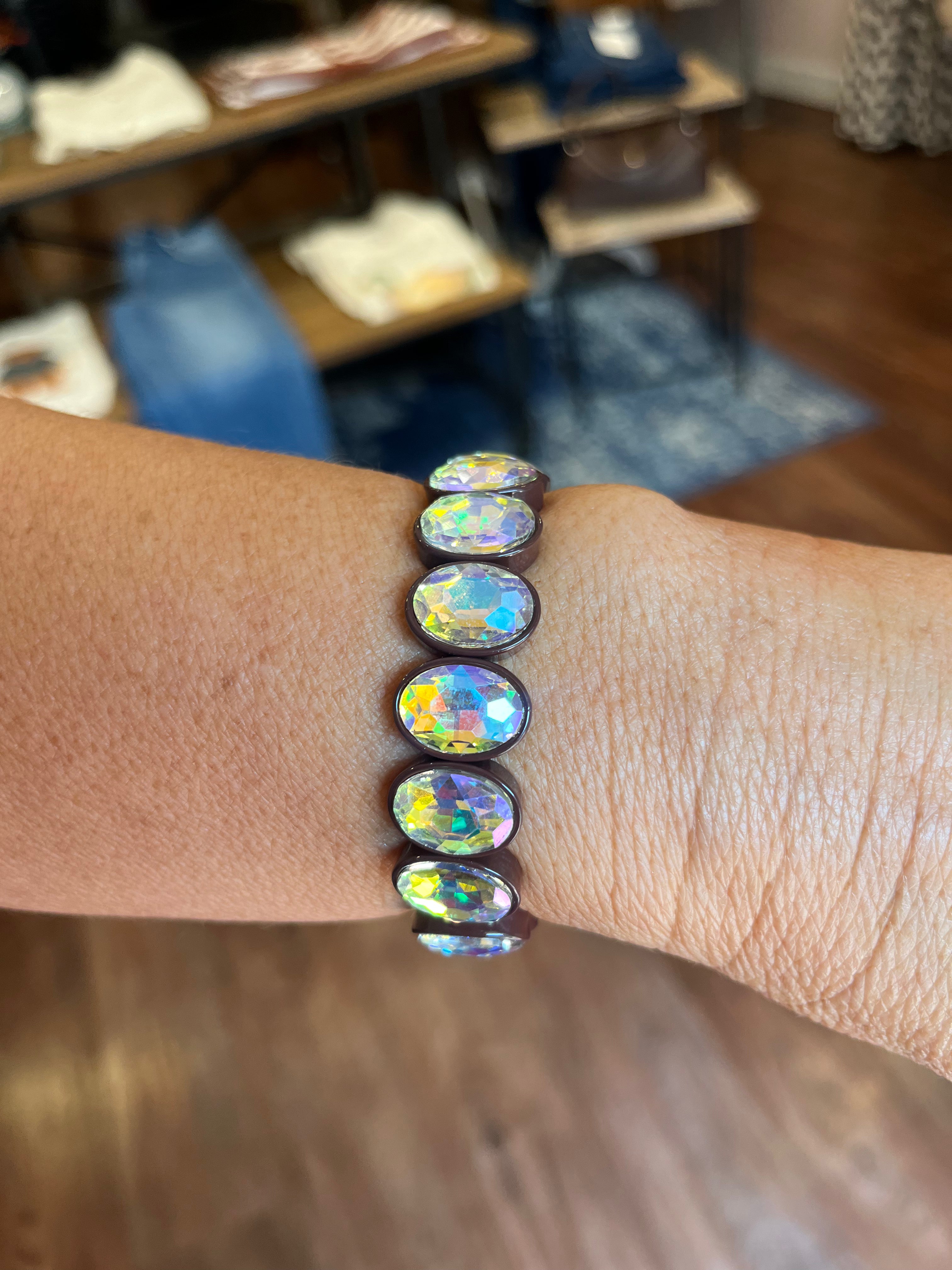 A close-up of a person's wrist wearing a dark brown elastic bracelet with seven oval iridescent cabochon gemstones that shimmer with rainbow colors, set against a blurred indoor background.