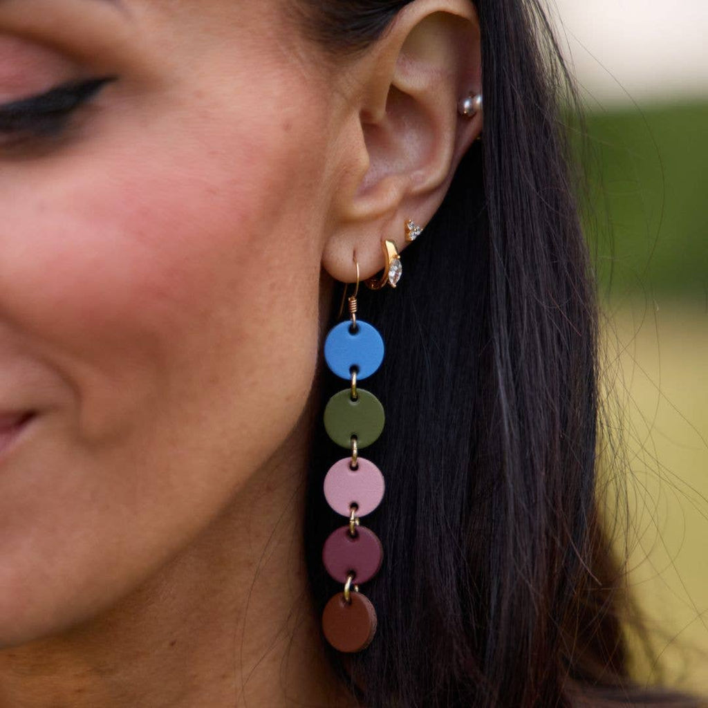 A close-up of a woman's ear wearing a long dangling earring made of stacked circular discs in blue, olive green, pink, mauve, and brown with small gold-tone rings and hooks.