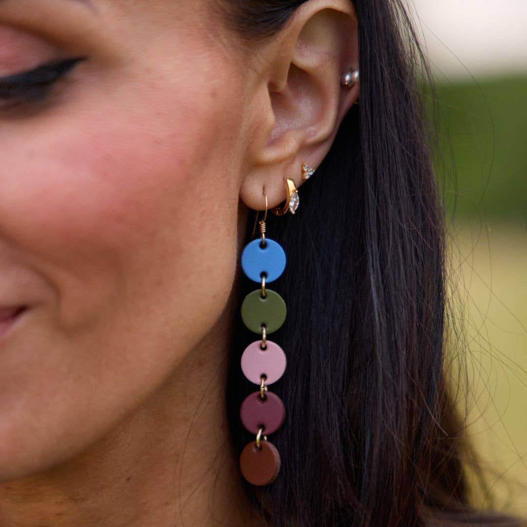 A close-up of a woman's ear wearing a long dangling earring made of stacked circular discs in blue, olive green, pink, mauve, and brown with small gold-tone rings and hooks.