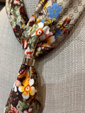 A close-up of a patterned silk scarf tied in a knot with a gold ring, displayed on a textured beige fabric mannequin. The scarf features a floral print with white, yellow, blue, and red flowers on a beige background with black monogram-like patterns.