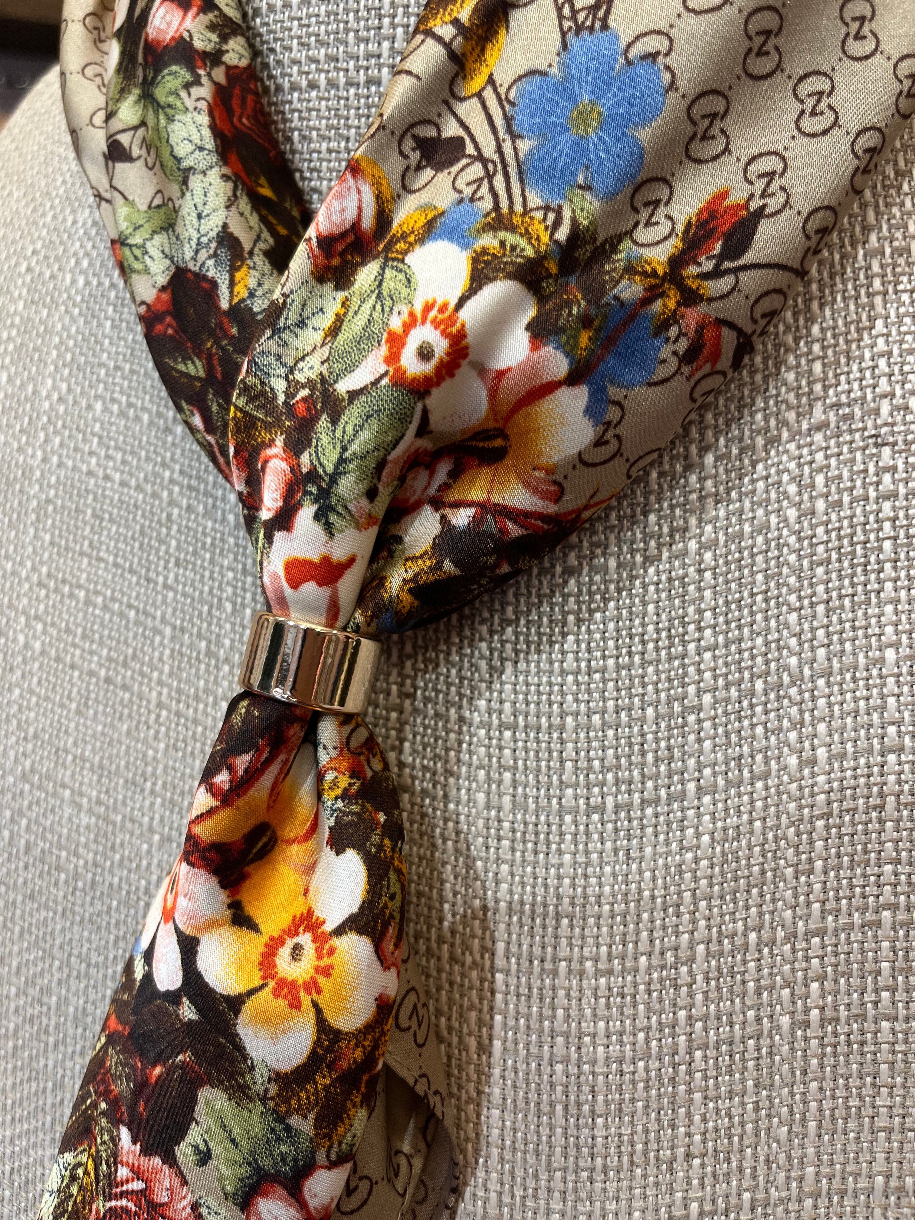 A close-up of a patterned silk scarf tied in a knot with a gold ring, displayed on a textured beige fabric mannequin. The scarf features a floral print with white, yellow, blue, and red flowers on a beige background with black monogram-like patterns.