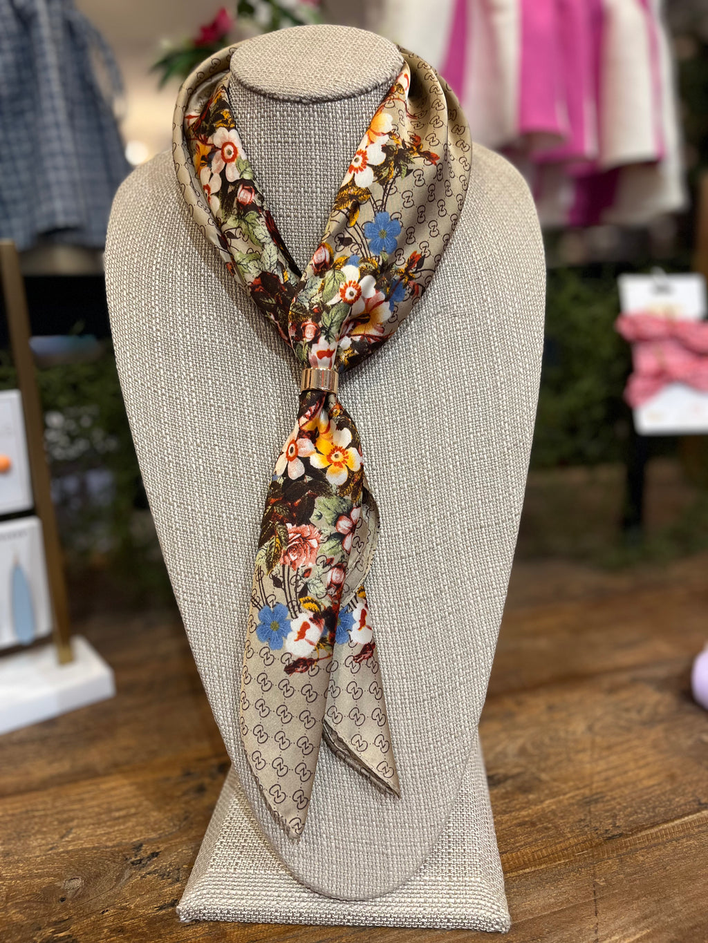 A patterned silk scarf with a floral print and geometric border, styled on a beige fabric mannequin bust in a retail store setting.