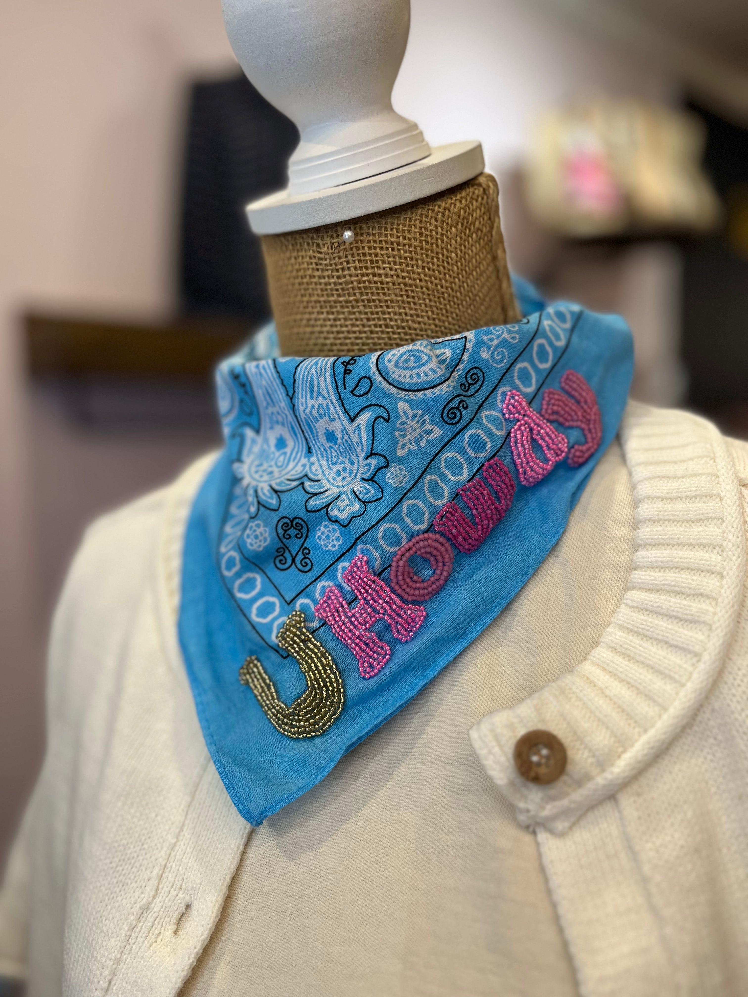 A bright blue bandana with white paisley patterns and pink embroidered text reading 'HOWDY' along with a gold horseshoe design, displayed on a cream-colored sweater dress form.