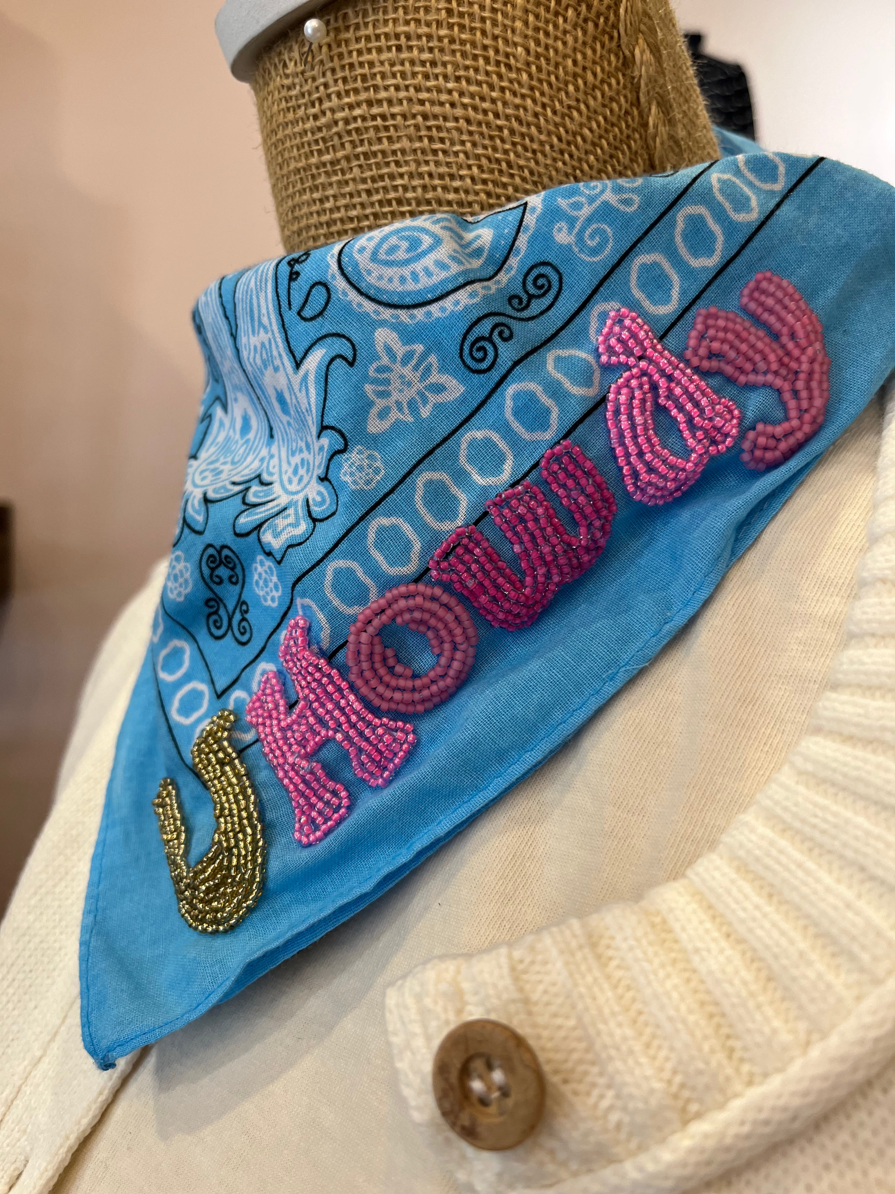 A blue bandana-style neck scarf with white paisley patterns and pink beaded lettering that reads "HOWDY," displayed on a cream knit sweater dress or mannequin torso.