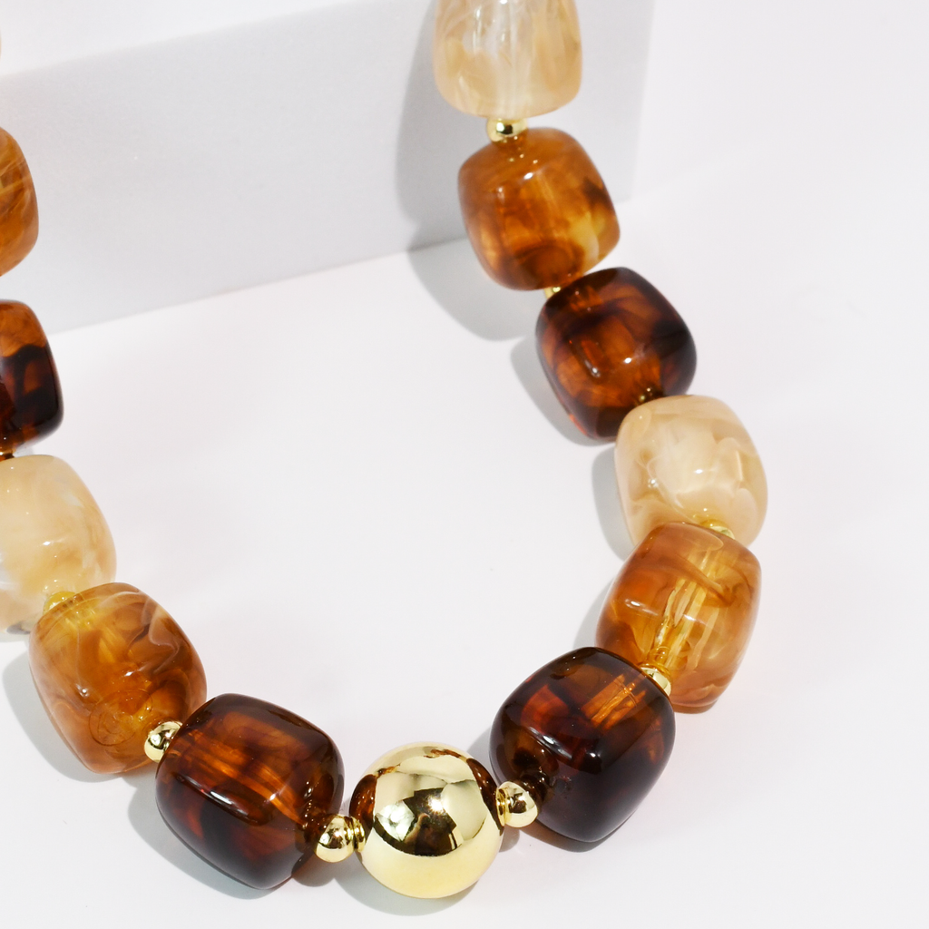 A beaded necklace with chunky, rounded translucent beads in amber, honey, and cream tones, accented by a shiny gold central bead and small gold spacer rings, displayed on a clean white surface.