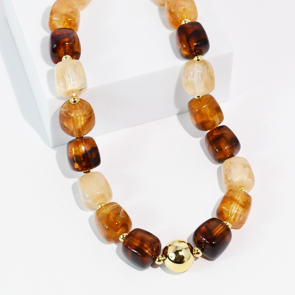 A beaded necklace with alternating translucent amber-brown and cream beads, accented by small gold spacers and a larger polished gold bead at the center, displayed on a white block against a white background.