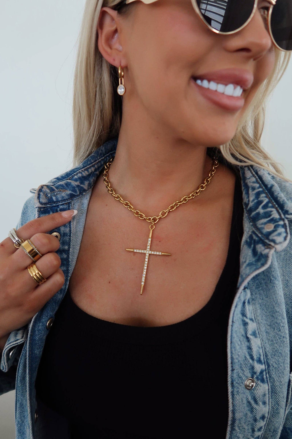Woman wearing a gold necklace with a cross pendant, denim jacket, and sunglasses.