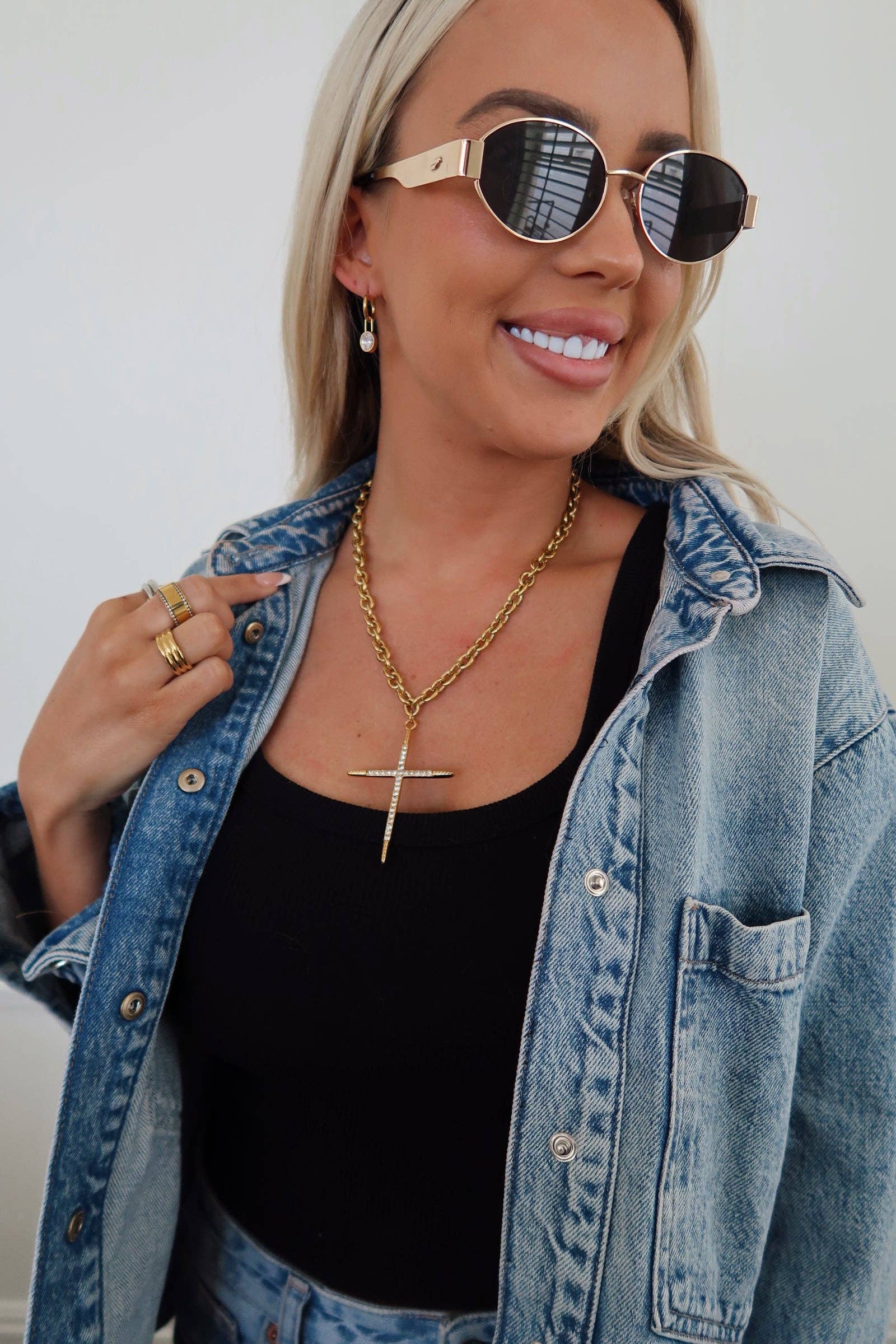 Woman wearing sunglasses, a denim jacket, and black top with gold jewelry.