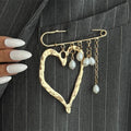 A gold heart-shaped brooch pinned to a gray pinstripe blazer pocket, featuring dangling pearl-like beads and chain accents. A hand with long white manicured nails is visible on the left side.
