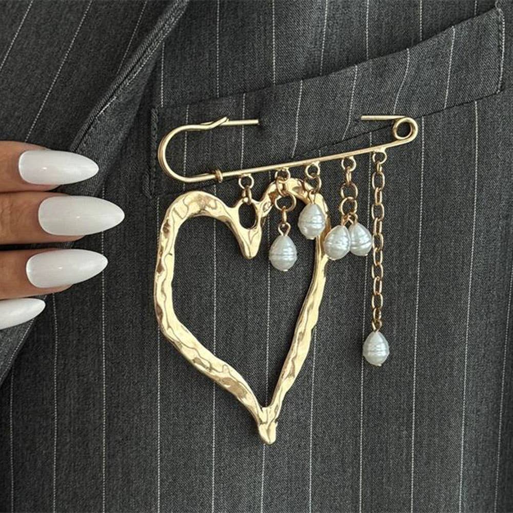 A gold heart-shaped brooch pinned to a gray pinstripe blazer pocket, featuring dangling pearl-like beads and chain accents. A hand with long white manicured nails is visible on the left side.