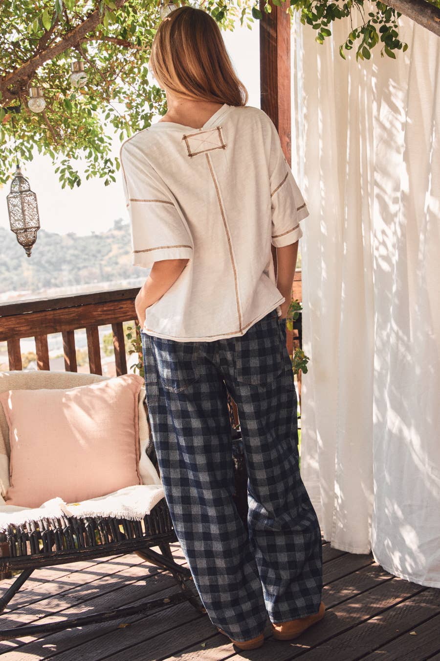 Person standing on a wooden deck with a scenic view, wearing a white top and plaid pants.