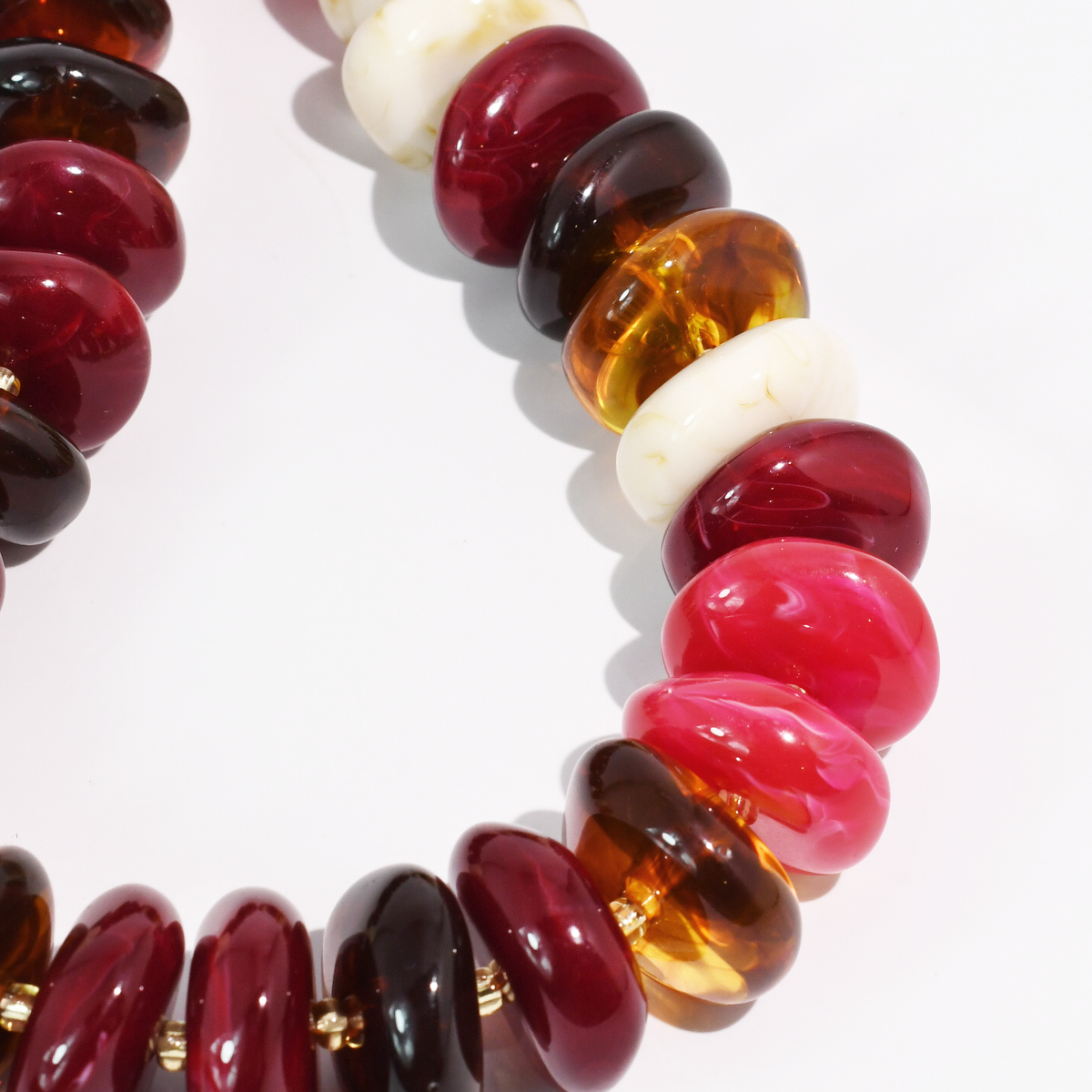 A close-up of a beaded necklace featuring glossy, polished disc-shaped beads in deep burgundy, translucent amber, bright pink, and off-white, arranged in a circle on a clean white background.