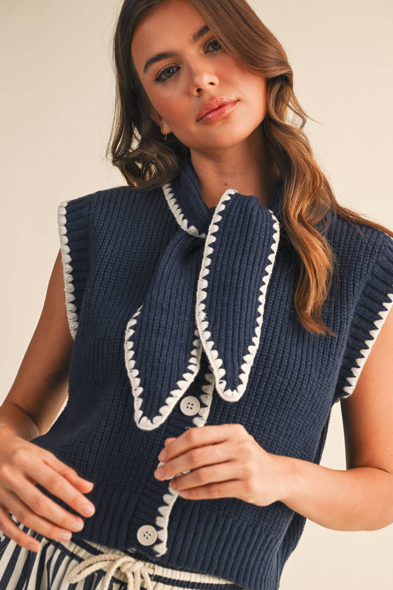 Woman wearing a navy blue knitted vest with white trim and buttons on a beige background