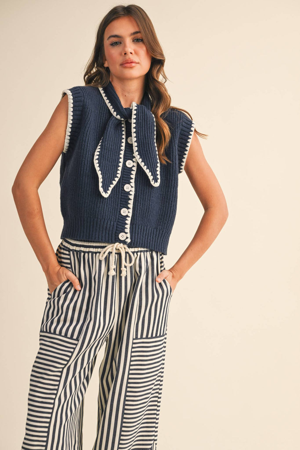 Woman wearing a navy blue sleeveless knitted top with white buttons and striped pants on a beige background