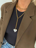 Person wearing a gold chain necklace with a heart pendant, brown blazer, black top, and blue jeans.