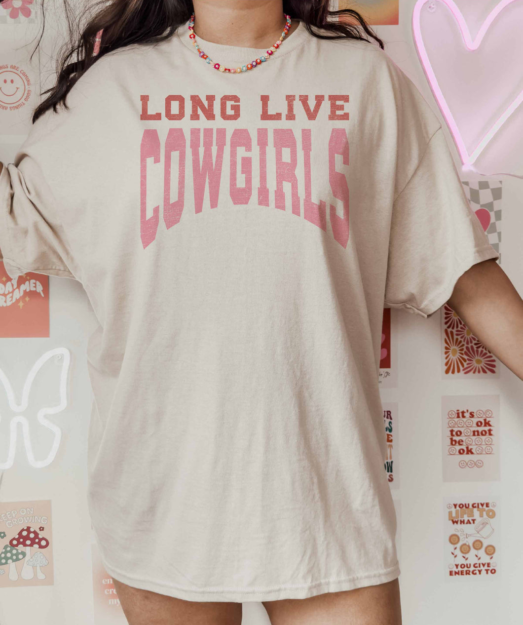 Person wearing a beige t-shirt with 'LONG LIVE COWGIRLS' text in pink, standing against a colorful wall.
